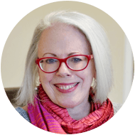 Portrait of a smiling older woman with white hair, wearing red glasses and a vibrant pink and purple scarf. She appears friendly and approachable, ready for professional residencies.