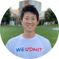 A smiling Asian man wearing a white t-shirt with the text "WEINDMIT" standing in a park with city buildings in the background.