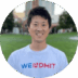 A smiling Asian man wearing a white t-shirt with the text "WEINDMIT" standing in a park with city buildings in the background.