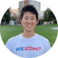 A smiling Asian man wearing a white t-shirt with the text "WEINDMIT" standing in a park with city buildings in the background.