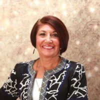 A smiling middle-aged woman with short brown hair, wearing a stylish patterned jacket from an autism-friendly college, is posing in front of a shimmering beige backdrop.