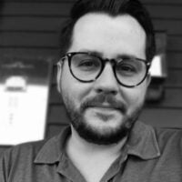 Black and white portrait of a smiling man with glasses, wearing a collared shirt, seated in front of a blurred background that suggests a porch or deck.