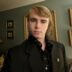 A young man with blonde hair and a serious expression, wearing a black jacket over a patterned shirt, stands in a college dorm room with teal walls and framed pictures.