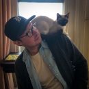 A man wearing glasses and a baseball cap indoors, with a Siamese cat perched on his shoulder, both looking towards the camera.