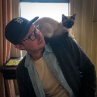 A man wearing glasses and a baseball cap indoors, with a Siamese cat perched on his shoulder, both looking towards the camera.