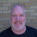 A smiling bald man with a beard, wearing a black t-shirt and glasses perched on his head, standing in front of a brick wall.