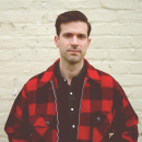 A man in a red and black plaid jacket stands in front of a white brick wall, looking directly at the camera with a neutral expression.