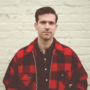 A man in a red and black plaid jacket stands in front of a white brick wall, looking directly at the camera with a neutral expression.
