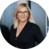 A professional woman with blonde hair wearing glasses and a dark blazer smiles slightly, standing against a blurred green background. Her portrait is encircled by a soft, round frame.
