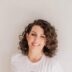 A woman with curly hair smiling at the camera, wearing a white T-shirt against a plain light background.