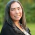 Portrait of a smiling Asian woman with long gray hair, wearing a black blazer and a pearl necklace, outdoors with soft-focus greenery in the background, symbolizing resilience in combating healthcare disparities.