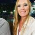 A woman with blonde hair smiling at the camera, wearing a white shirt, with a night-time cityscape illuminated in the background.