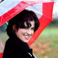 A smiling woman with short dark hair, holding a red and transparent umbrella, is photographed outside with green blurred background.