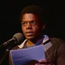 A man with glasses reads from a paper at a microphone on a dimly lit stage, wearing a sweater vest over a shirt.