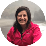 Woman in a pink jacket smiling at the camera with a misty and wooded river or lake landscape in the background.