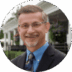 A professional middle-aged man with glasses smiling, wearing a blue shirt, tie, and suit jacket, with a blurred house and greenery in the background.