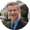 A professional middle-aged man with glasses smiling, wearing a blue shirt, tie, and suit jacket, with a blurred house and greenery in the background.