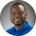 A smiling man with a beard, wearing a blue polo shirt, set against a blurred greenish background. His expression is friendly and approachable.