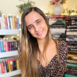 A woman with long, ombré hair and a floral dress smiling warmly, surrounded by shelves filled with colorful books.