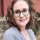 A portrait of a smiling woman with curly hair wearing glasses and a gray t-shirt, standing against a blurred background of a red wall and greenery.