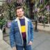 A man smiling at the camera, wearing a denim jacket with a shearling collar and a multicolored sweater, standing in front of a floral trellis.