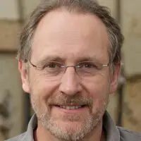 Close-up portrait of a smiling middle-aged man with glasses, short graying hair, and a beard. He is wearing a casual shirt, and the background is softly blurred.