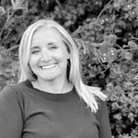 Black and white portrait of a smiling woman with blonde hair, wearing earrings and a dark sweater, standing in front of lush foliage.