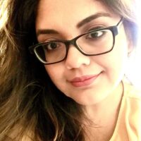 A close-up portrait of a woman with dark hair wearing black-rimmed glasses and a yellow top, looking at the camera with a subtle smile.