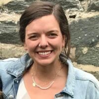 A smiling woman with shoulder-length brown hair, wearing a denim jacket and a pearl necklace, stands in front of a stone wall.