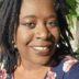 A close-up portrait of a smiling woman with dreadlocks and sunlit face, wearing earrings and a floral top, framed by green leaves in the background.