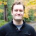 Man in a black sweater smiling at the camera, with autumn trees in soft focus in the background.