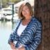 A woman with short hair wearing a striped blue and white cardigan leans against a wooden post with a marina in the background.