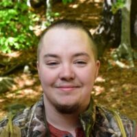 A smiling person with a buzz cut wearing a camouflage jacket over a red shirt, posing in front of blurred greenery and trees.