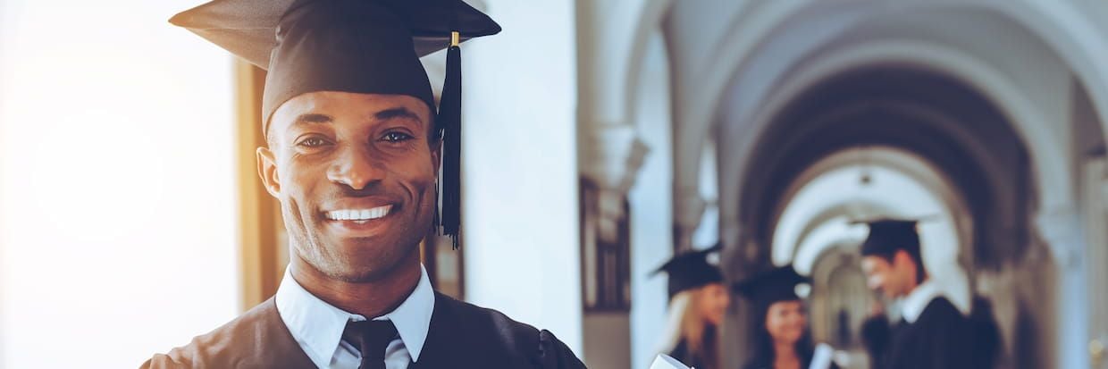 A happy graduate in a cap and gown smiling confidently in a university hallway, with other graduates conversing about scholarship resources in the background.