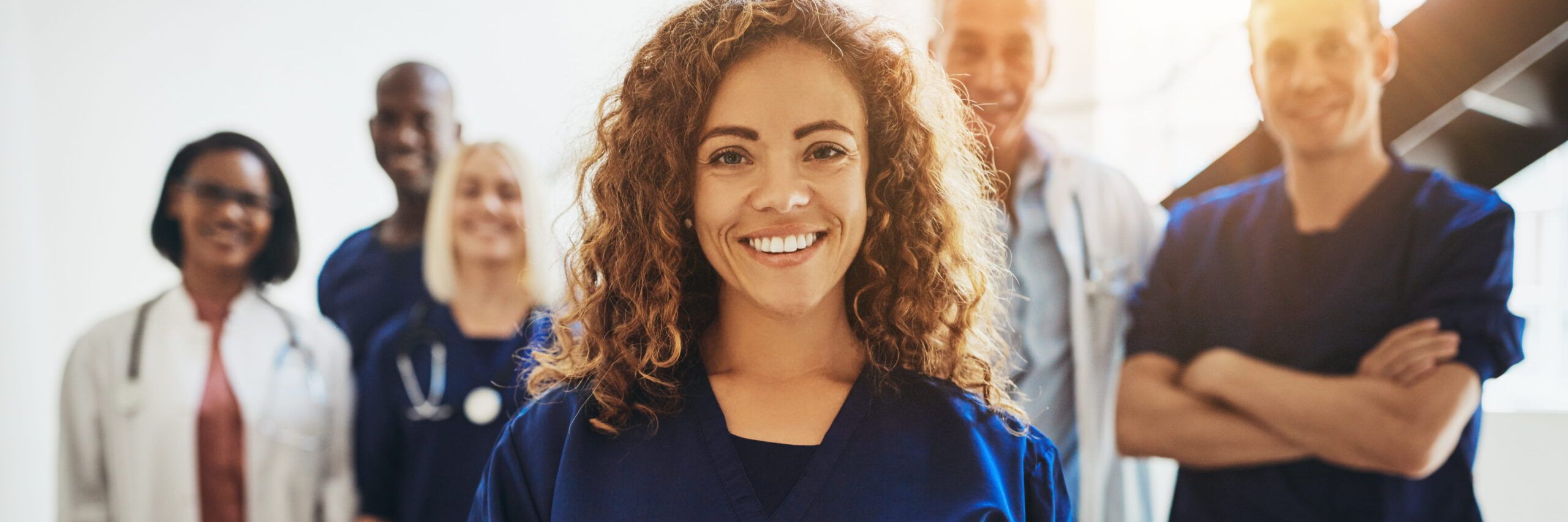 Smiling female healthcare worker in blue scrubs stands at the forefront, with a diverse group of medical professionals in various uniforms blurred in the background, all beneficiaries of education grants.