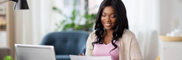 A smiling black woman with a PhD in a pink top reviews documents at her desk, with a laptop and lamp nearby in a well-lit, cozy office setting.