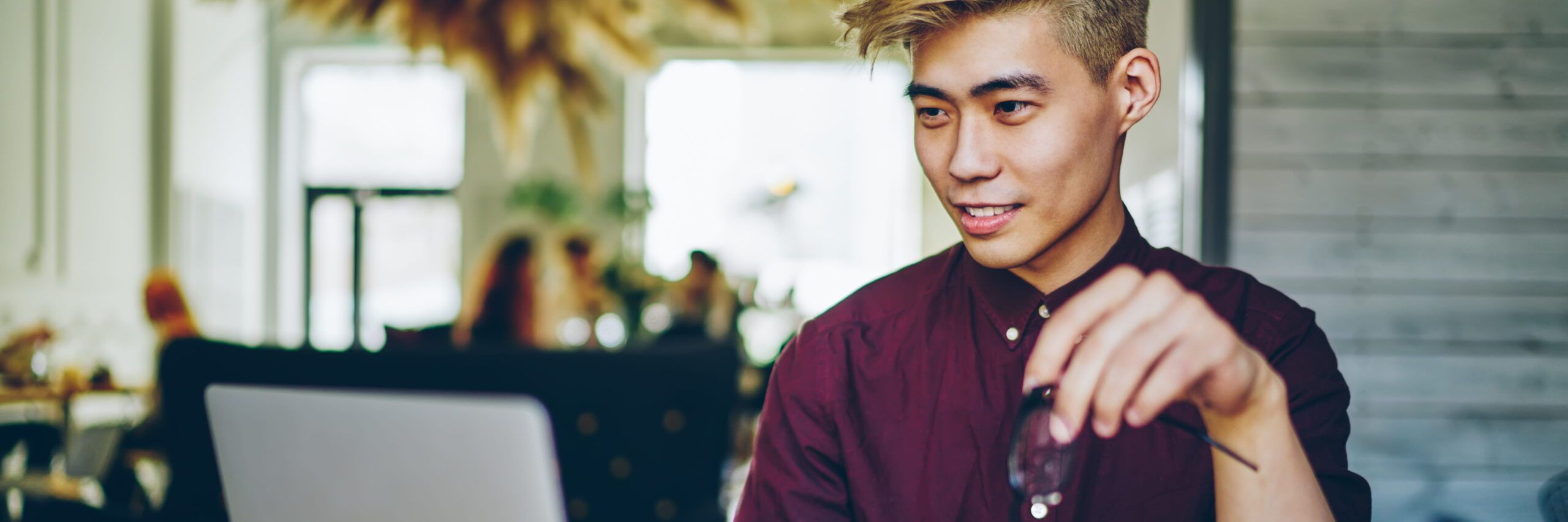 Young Asian man in a burgundy shirt working on a laptop in a modern café, smiling slightly as he applies for scholarships online.