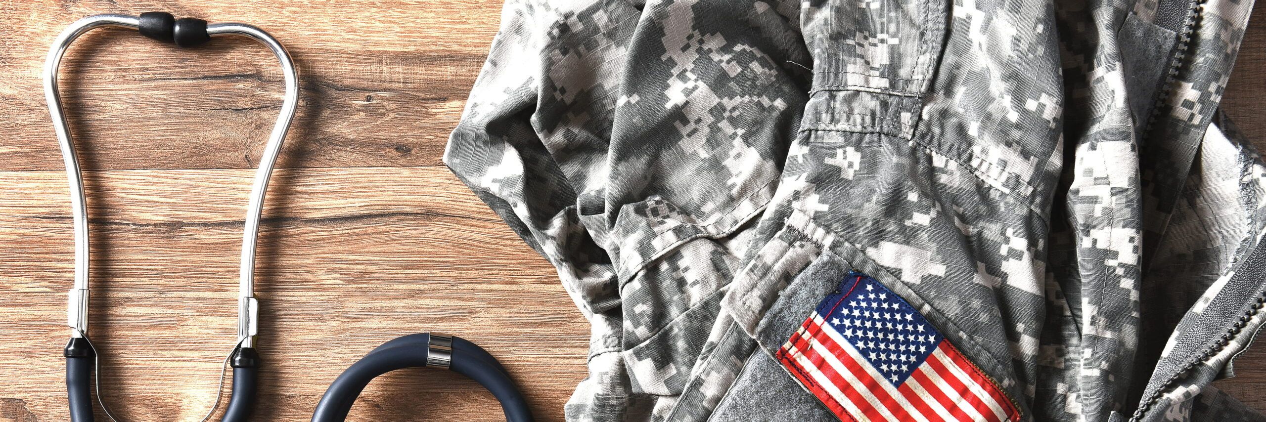 A black stethoscope lying next to a folded military uniform with a U.S. flag patch on a wooden surface, symbolizing veterans' medical services.