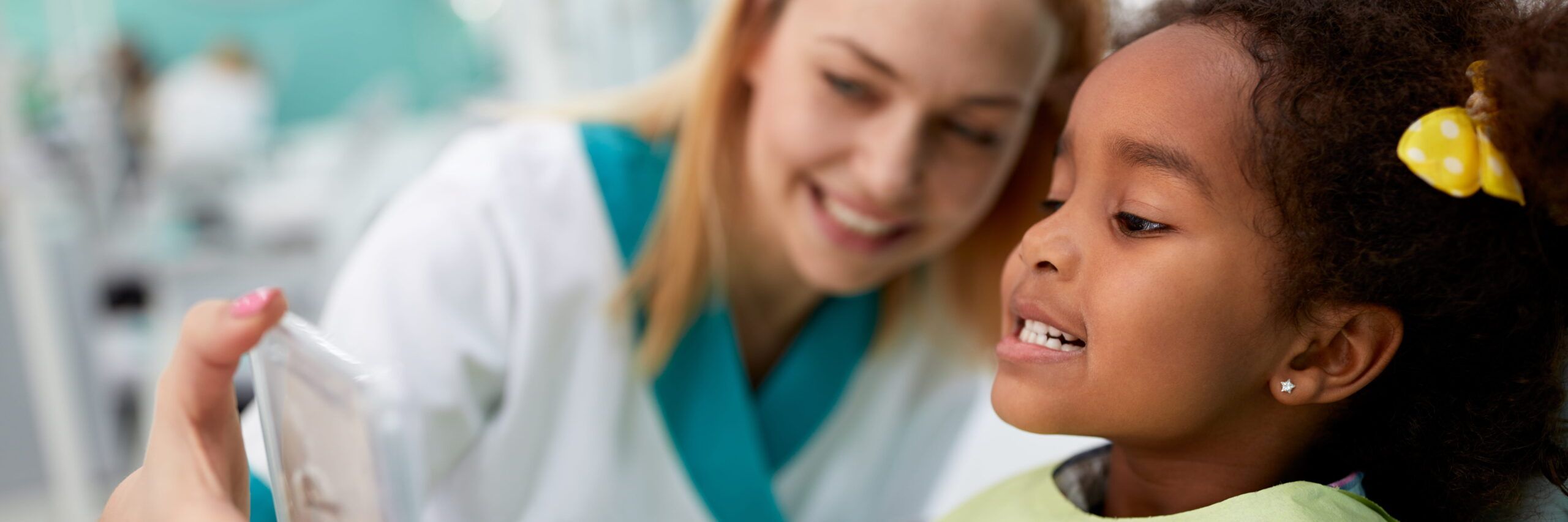 A young dentist, a white woman, shows a dental x-ray to a black girl with curly hair, who looks interested. They are in a bright, clean dental office with her dental assistant nearby.