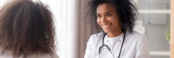 A smiling female doctor with a stethoscope around her neck talking to a patient about women's health in a clinic, viewed from the patient's back.