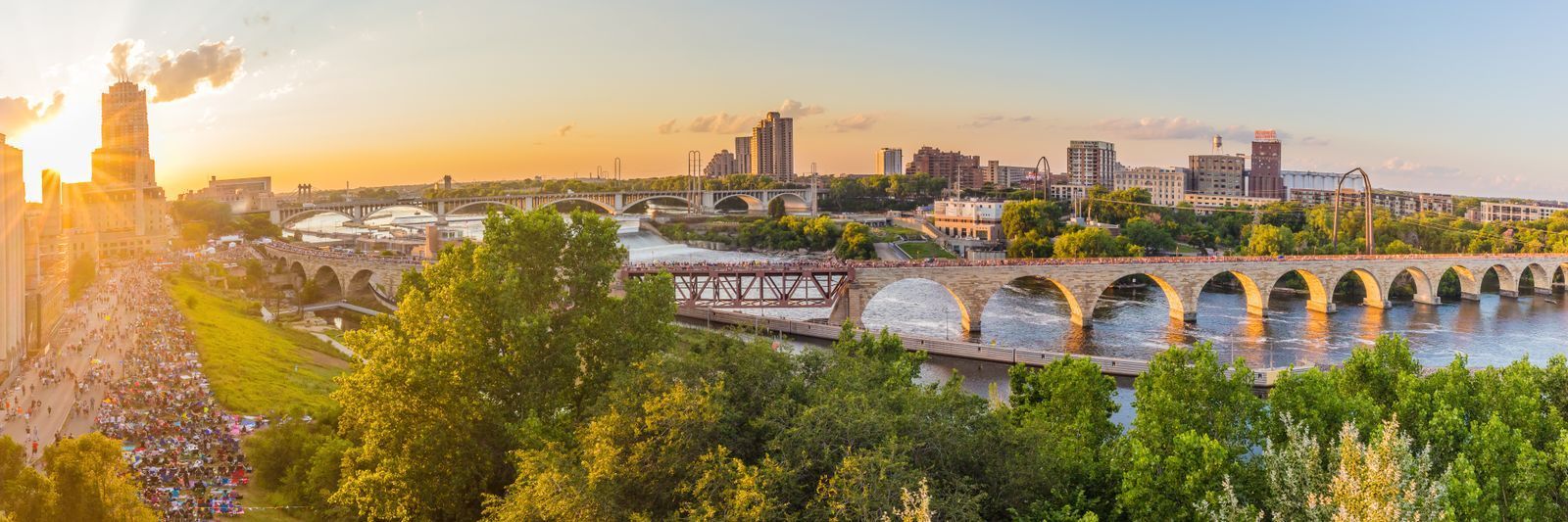 Panoramic view of a cityscape at sunset featuring a river with multiple bridges, a bustling promenade, and modern buildings under a wide sky with scattered clouds.