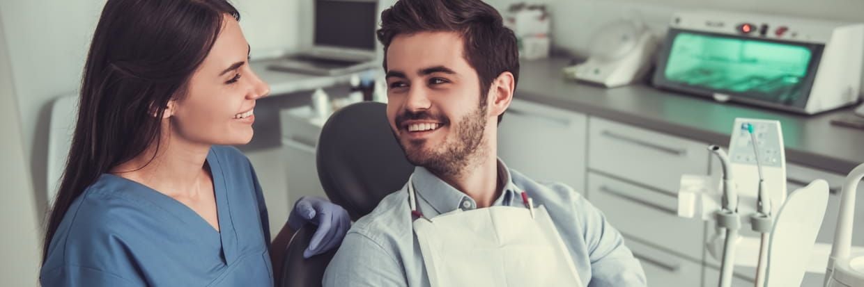 A female dentist and a male patient smiling at each other in a dental clinic with dental equipment and dental education materials visible in the background.