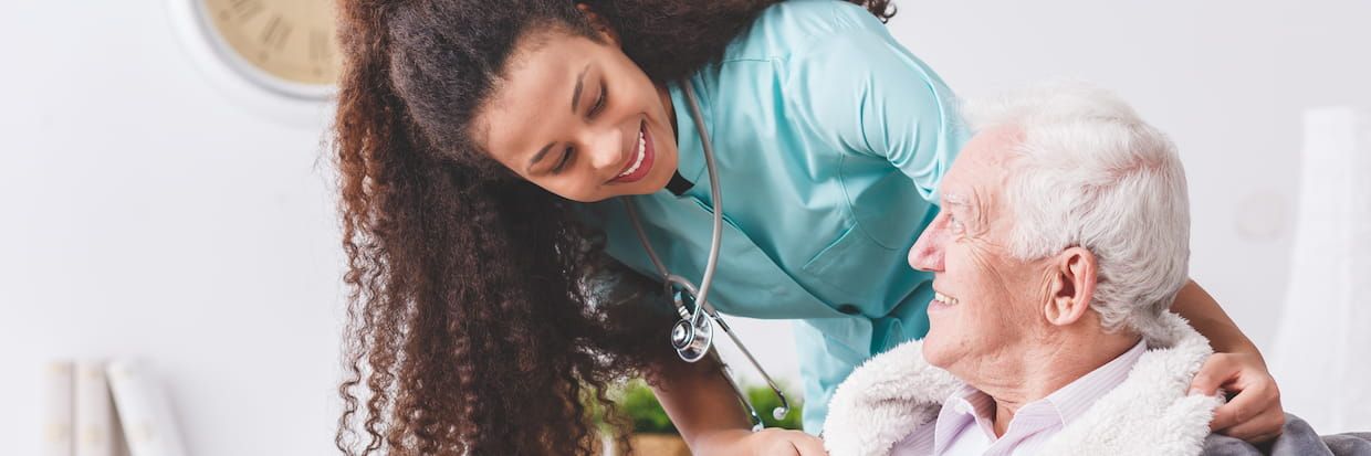 A smiling female nurse with curly hair attending to an elderly man in a wheelchair, both looking happy and engaged in a warm conversation about his Home Health Aide certification.