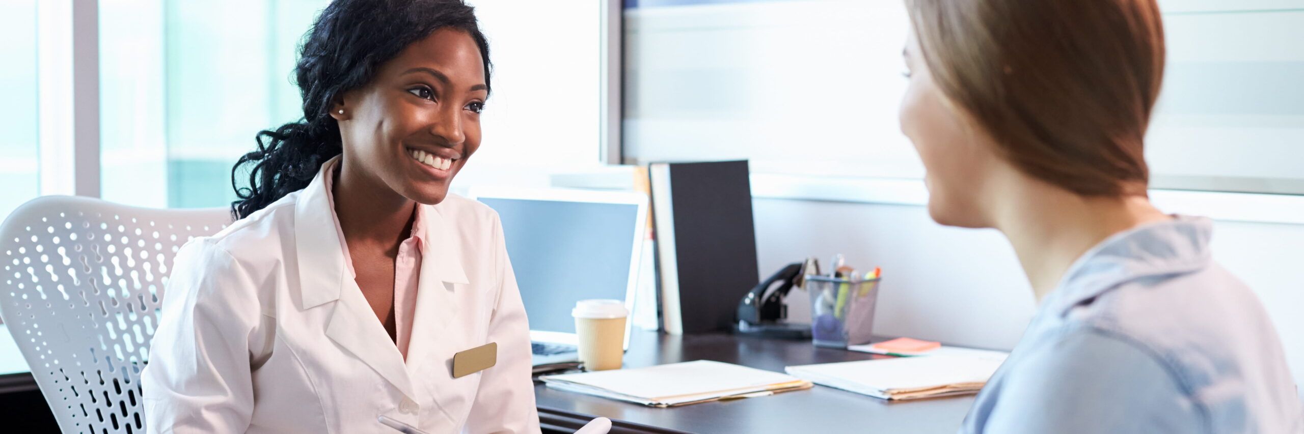 A female doctor with a name tag, smiling at a female patient during a consultation about medical billing in a bright office setting.