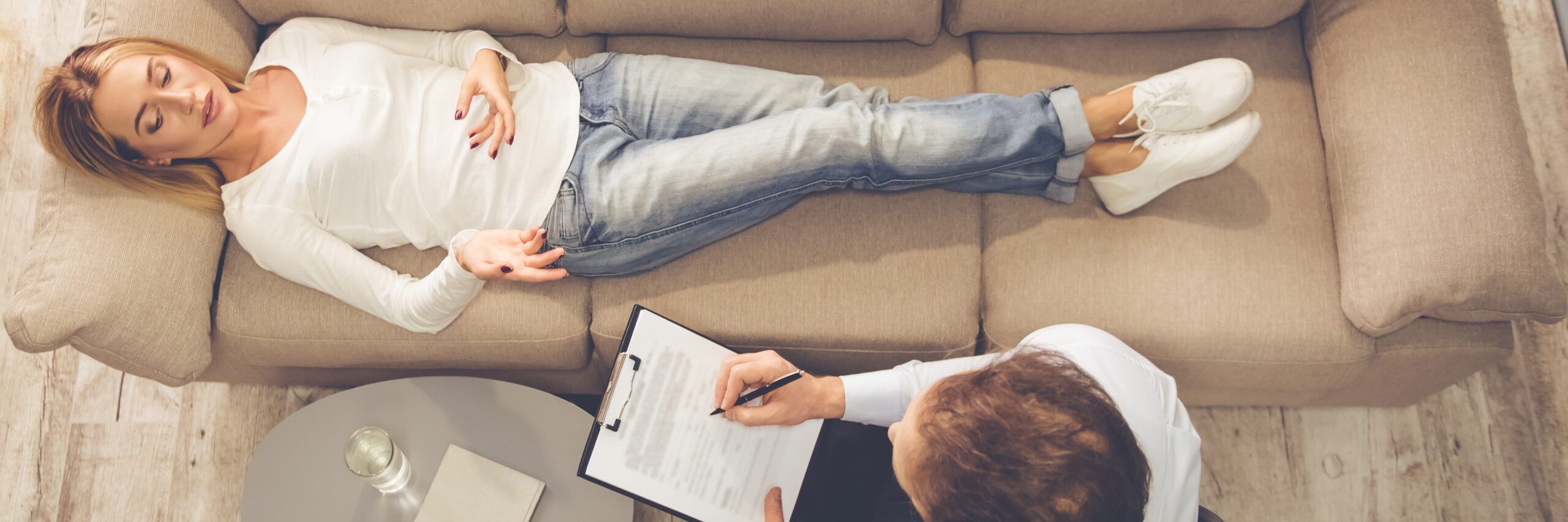 A woman reclines with closed eyes on a beige sofa, while a man sitting beside her writes on a clipboard, suggesting a psychiatric therapy session.