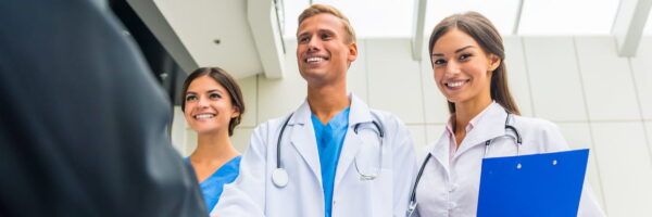 Three healthcare professionals, two women and one man, smiling and interacting with a patient or colleague, acing admissions interviews, wearing white lab coats and stethoscopes in a bright clinical setting.