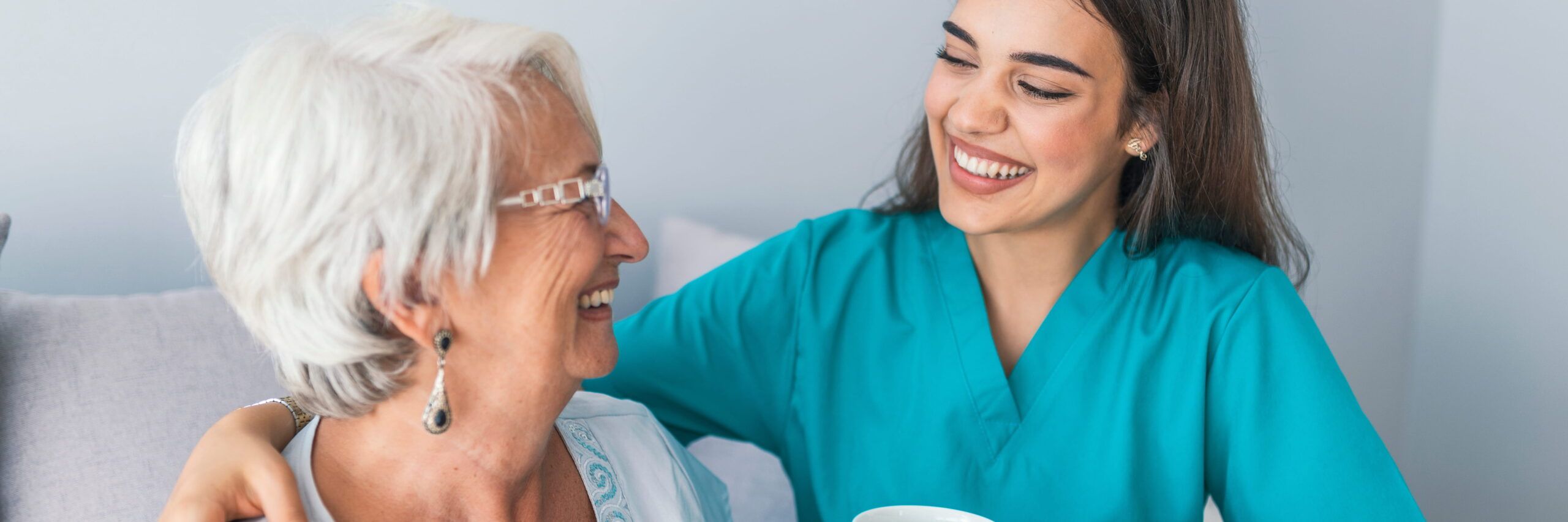 A cheerful caregiver in teal scrubs smiles at an elderly white-haired woman wearing glasses, both sharing a joyful moment on a gray sofa. The caregiver, a recent college graduate, holds a white cup.