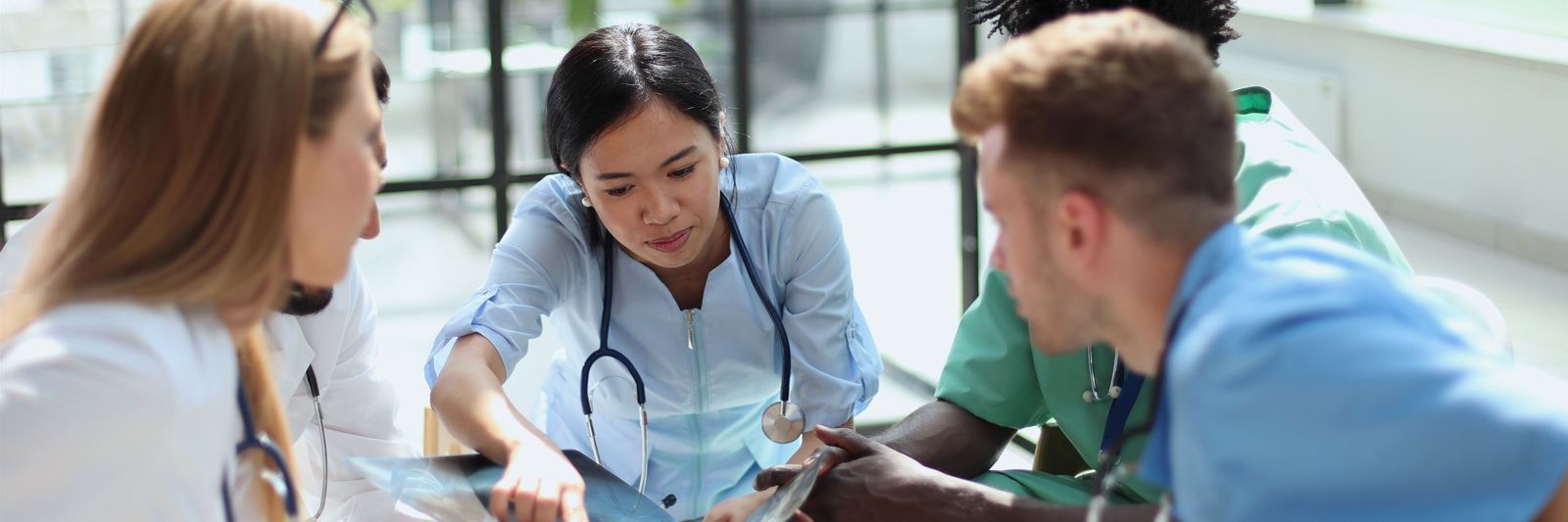 A diverse group of medical professionals, including an asian female doctor and three colleagues, actively discussing over documents in a well-lit modern healthcare setting.