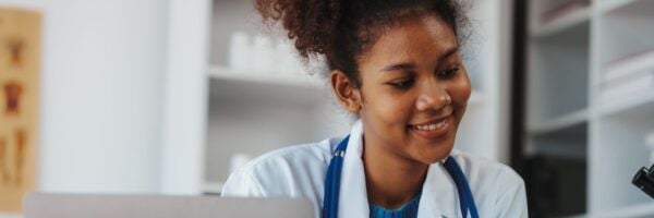 A young female doctor with curly hair, wearing a stethoscope, smiles while looking at her laptop in a modern office setting.