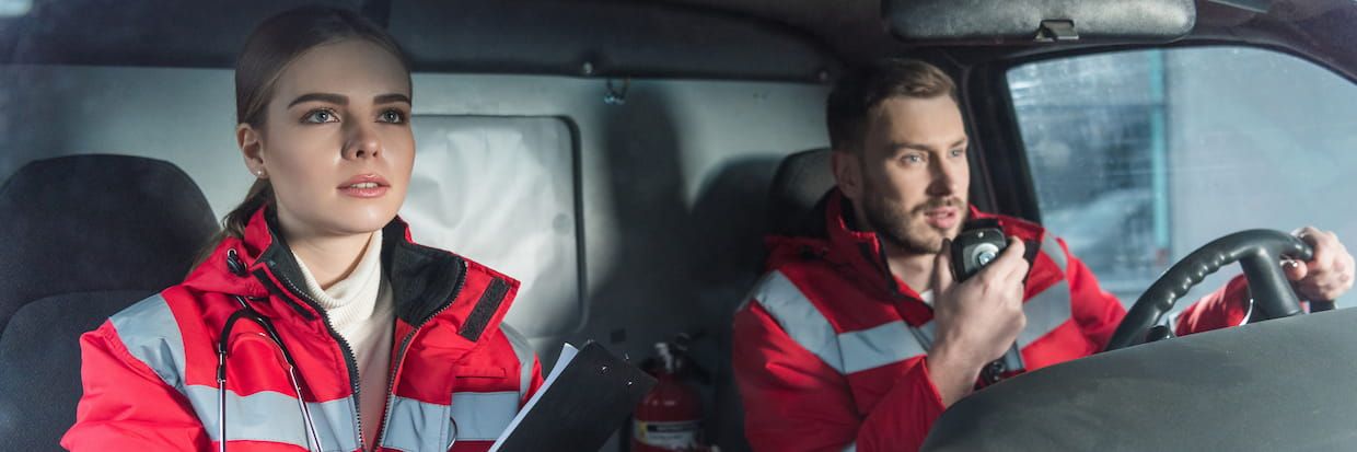 Two paramedics in an ambulance, a focused male driver speaking into a radio and a female with a clipboard looking intently out the window, both part of an emergency medical services team.